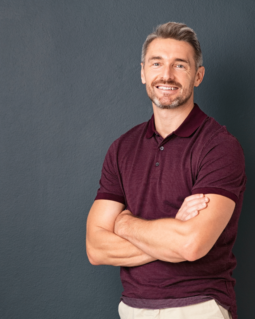 Smiling man with crossed arms, wearing a maroon polo shirt, against a gray background, representing Prime Vitality's focus on men's health and wellness services.